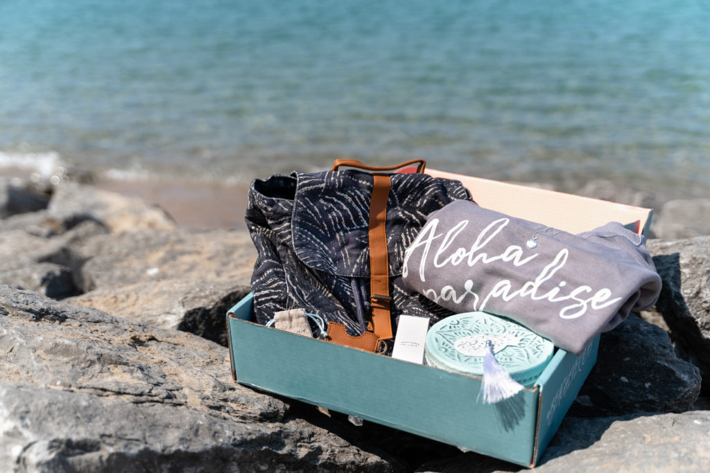 Box of beach inspired products on a rock near water.